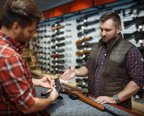 Gun store owner assisting a customer in selecting a handgun, with rifles displayed on the wall.