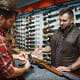 Gun store owner assisting a customer in selecting a handgun, with rifles displayed on the wall.