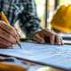 Close-up of a contractor signing a construction agreement with a yellow hard hat in the background.